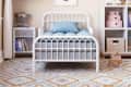Child's bedroom with white metal bed, blue bedding, geometric rug, and shelves with toys and books.