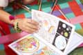 Child coloring a comic book on a colorful mat with colored pencils scattered around.