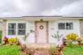 White house with light pink front door. in front of cloudy sky. Green grass and yellowish reddish bushes out front