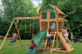 Children playing on a wooden playset with swings, slide, and climbing wall in a grassy backyard.