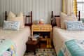 Two twin beds with patterned quilts, decorative pillows, a rattan nightstand, and yellow rain boots on a rug.