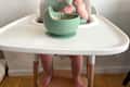 A baby in a high chair playing with a green bowl of food, with bare feet visible on the wooden footrest.