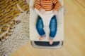 A child in a striped orange sweater sits in a cream-colored baby bouncer on a wooden floor, with a patterned rug nearby.