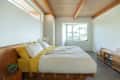 Minimalist bedroom with wooden beams, a bed with yellow sheets, white quilt, and a wooden bench with folded towels and books.
