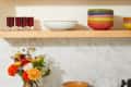 Colorful ceramic bowls stacked on wooden shelves with flowers in vases and red glasses on a marble countertop.