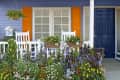 Front porch with white rocking chairs, vibrant flowers, orange shutters, and a blue door.