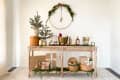 Festive holiday bar setup with a wooden table, greenery, wrapped gifts, a wreath, and assorted drinks and decor.