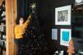 Woman in yellow sweater decorates black Christmas tree with gold star in cozy living room.