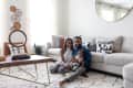 Family sitting on a patterned rug in a living room with a beige sectional sofa, geometric pillows, and a round mirror.