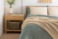Cozy bedroom with a wooden bed, green bedding, beige pillows, a nightstand with a vase, and a woven wall hanging.