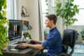 Man in blue shirt working at a desk with computer, surrounded by plants in a bright room.