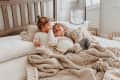 Two children in cozy pajamas on a bed with a plush toy, next to a humidifier on a nightstand.