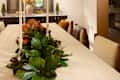 Dining room featuring a long table with a floral centerpiece, white candles, and upholstered chairs, illuminated by a modern chandelier.