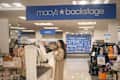 A woman in a beige trench coat holds a white item in a Macy's Backstage store, surrounded by clothing racks and festive decor.
