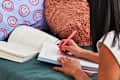 closeup shot of girl laying on a bed with teal sheets, smiley face pillowcase  and a furry circled pillow writing in a notebook with a pink pen and a text book in front of her