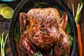 Roasted turkey in a black pan with herbs, surrounded by carrots, lemon halves, green beans, and a bowl of mixed vegetables.