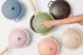 Assorted colorful pots with wooden handles, including blue, green, pink, and beige, arranged on a light surface.