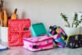 Colorful lunch bags and containers on a kitchen counter with wooden utensils and potted plants in the background.