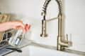 Hand holding a spray bottle near a stainless steel kitchen faucet.