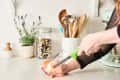 Hand lighting a candle in a glass jar on a kitchen counter with utensils and a plant in the background.