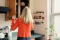 Woman in orange sweater organizing spices on kitchen shelves near a window with a potted plant.