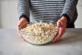 Person holding a glass bowl filled with freshly popped popcorn on a marble countertop.