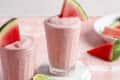 a pink, creamy, watermelon smoothie with a slice of watermelon as a garnish, and some slices of watermelon scattered around the glass.