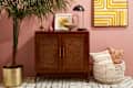 wide shot of entryway with palm tree in a golden planter, decorative pillows placed in cream woven basket, a framed piece on pink wall, and small brown cabinet with a frame, lamp, cream bowl, decorative gold bird and a pink vase on top of it