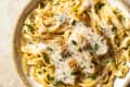 An overhead photo of a bowl of chicken fettucine Alfredo with grated parmesan cheese and parsley on top on a tan stone background.