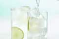Front view of two gin and tonics in high ball glasses, each with a lime in them and tonic being poured into the front glass.