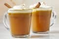 Head on view of two glass mugs of buttered apple cider topped with whipped cream and cinnamon sticks resting in the cups.