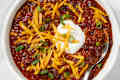 Overhead shot of chili in a white bowl, topped with sour cream, cheddar cheese and green onions - with a spoon resting in the bottom right of the bowl.