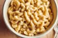 Creamy macaroni and cheese in a white bowl on a wooden table, with a red pot in the background.