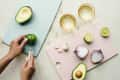 Hands slicing lime on a blue cutting board with avocado, garlic, onion, and lime on pink board, next to two glasses.