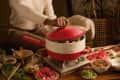 Person lifting lid of red hot pot on table with vegetables, mushrooms, and sliced meat, surrounded by bowls and chopsticks.