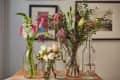Assorted flowers in glass bottles on a wooden table, with framed art in the background.