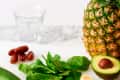 Pineapple, avocado, banana slices, spinach, zucchini, and dates on a marble surface with a glass.