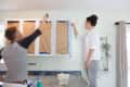 Two people painting kitchen cabinets white, using brushes and rollers, with protective paper covering the cabinet doors.