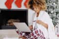 Woman in plaid pajamas shopping online with laptop and credit card, sitting by a Christmas tree and fireplace with stockings.