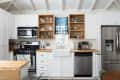 White kitchen with stainless steel appliances, open shelving, farmhouse sink, and blue curtain above the window.