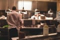 Chefs working in a busy restaurant kitchen with stacks of plates and hanging lights.