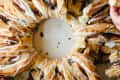 Cinnamon roll wreath with icing, being pulled apart by several hands on a white surface.