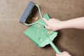 Hand sweeping crumbs into a green dustpan with a matching brush on a brown floor.