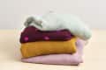 Stack of folded sweaters in pastel green, maroon with pink dots, mustard yellow, and light pink on a wooden surface.
