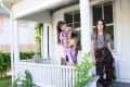 Two people on a porch, one holding a dog, surrounded by greenery and a white railing.