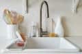 Kitchen sink with stainless steel faucet, white backsplash, dish soap, scrub brush, and utensils in a holder.