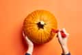 Hands carving a pumpkin with a red knife on an orange background.
