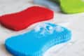 Red, blue, and green silicone scrubbers on a marble surface, with soap suds on the blue scrubber.