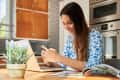 Woman in blue floral top using smartphone at kitchen table with laptop, next to a potted plant and open magazine.