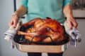 Person holding a roasted turkey in a metal roasting pan with striped towels.